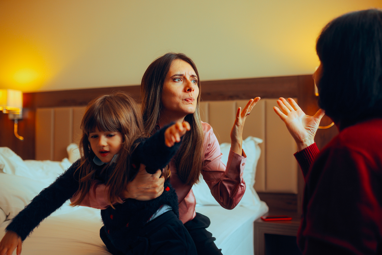 Warmly lit bedroom, frustrated mother holding upset child, facing seated adult, evening glow, documentary style, three people.