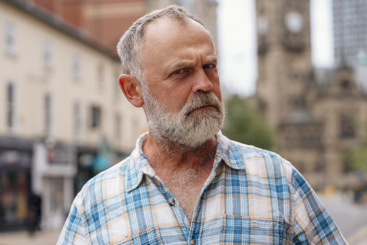 Town square scene, older man with gray beard foreground, shallow depth of field, daylight, editorial travel photo, people present.