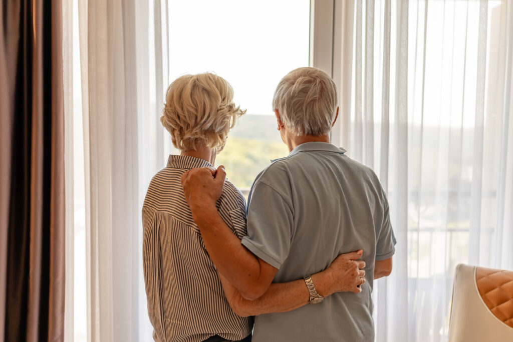 Bright living room, elderly couple arm in arm at window, backs to camera, soft daylight, documentary style, people present.