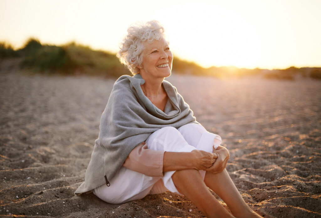 Beach, older woman with curly gray hair, seated wrapped in shawl, sunset glow, editorial travel photo, people.