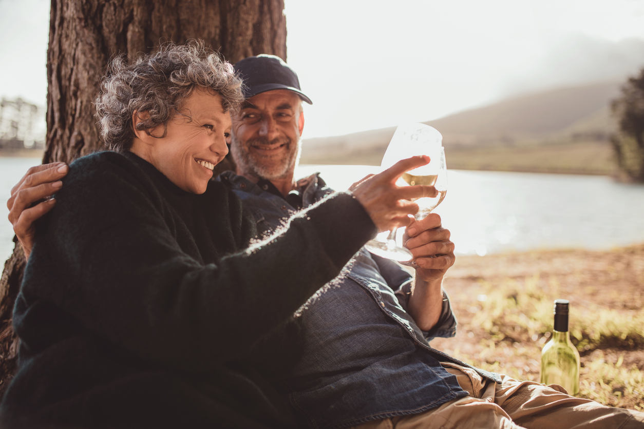 Lakeside tree shade, wine glasses foreground, couple close and smiling, soft golden hour light, editorial travel photo, people.