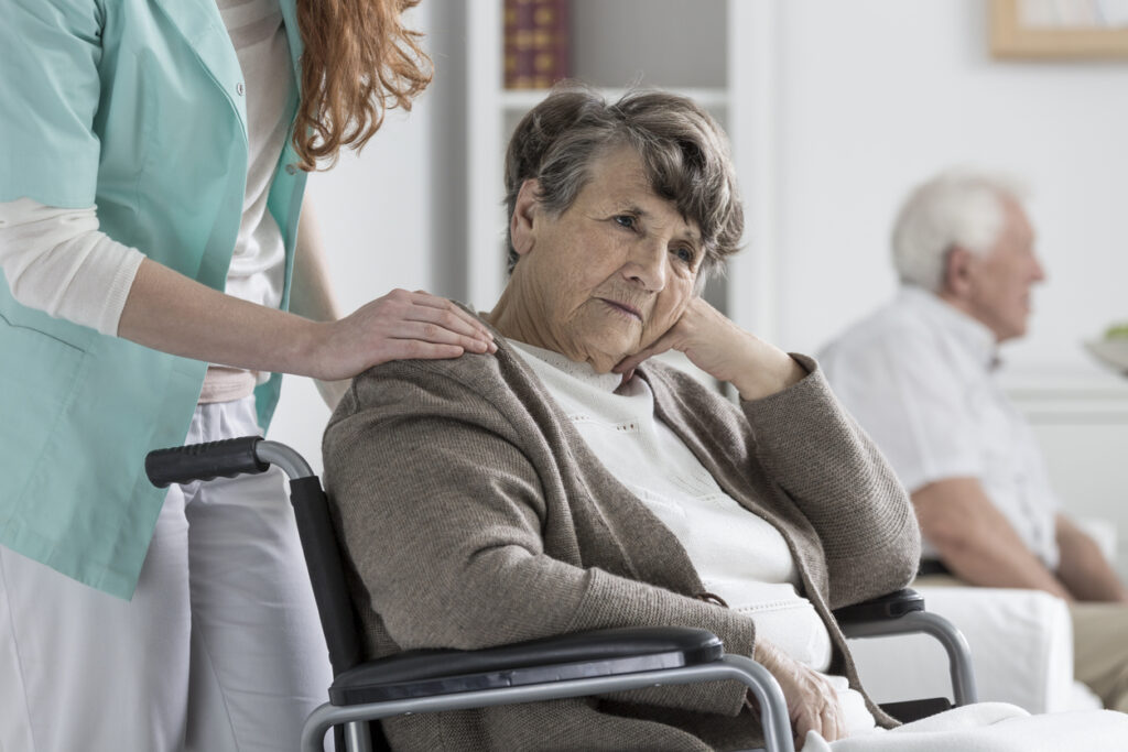Assisted living room, elderly woman in wheelchair foreground, caregiver hand on shoulder, morning light, documentary style, two people.