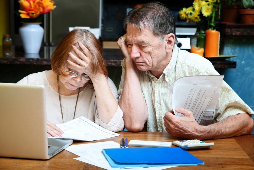 Kitchen table, worried elderly couple with bills and laptop, woman head in hand, daylight, documentary style, two people.