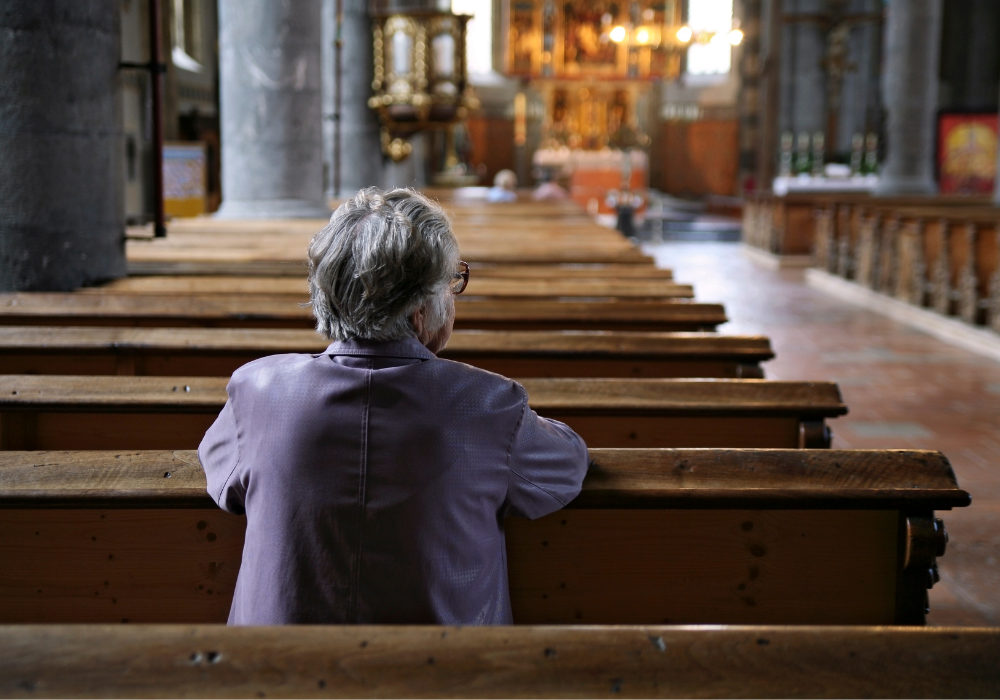 Large church interior, elderly person in wooden pew, facing ornate altar, soft morning light, editorial travel photo, solitary figure.