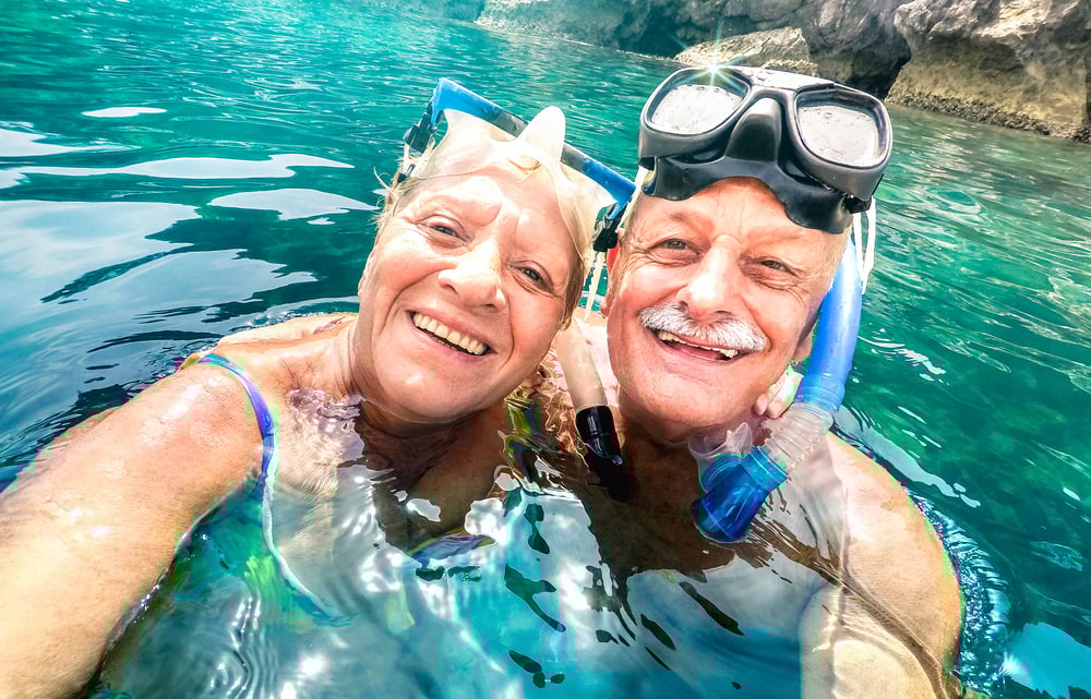 Tropical blue water, cheerful older couple selfie, mid-frame composition, midday sunlight, editorial travel photo, people.