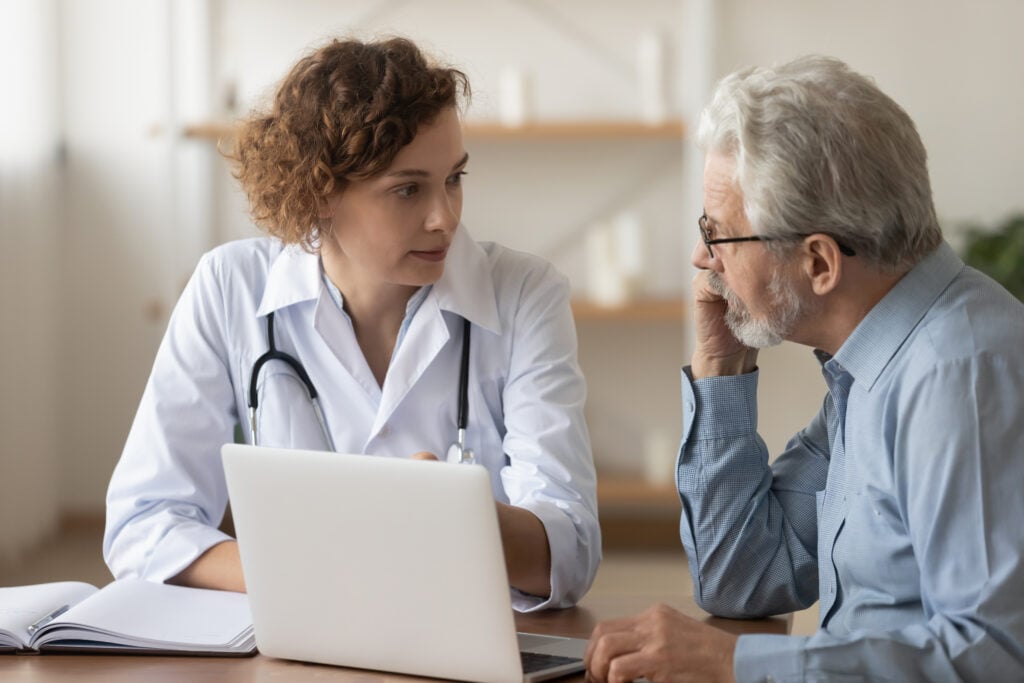 Medical office, doctor-patient discussion, seated at desk with laptop, daytime natural light, documentary style, two people.