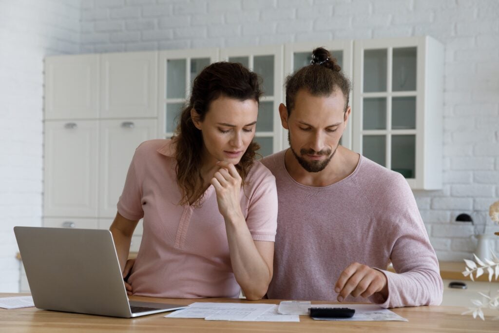Kitchen table, couple reviewing bills, documents and electronics scattered, morning light, documentary style image, people present.