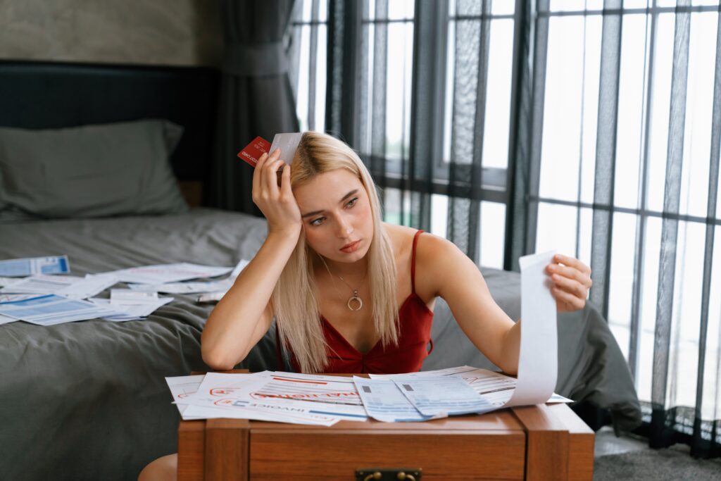 Cozy bedroom, stressed woman at cluttered table, bills and receipts foreground, afternoon light, documentary style, one person.