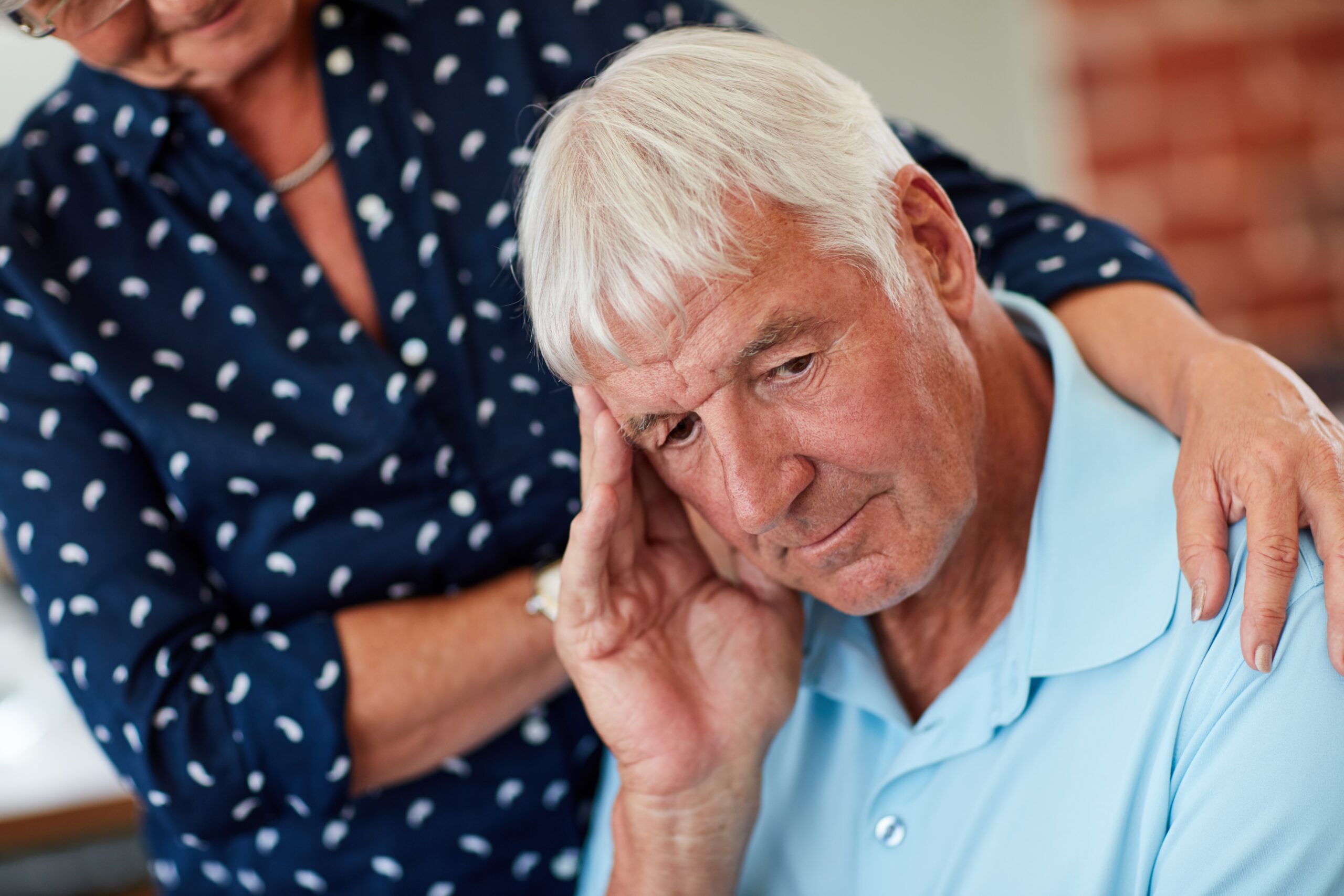 Living room, elderly man distressed foreground, woman beside him supportive, soft natural light, documentary style, two people.