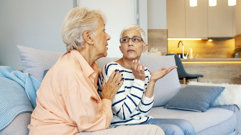 Living room, two older women in tense discussion, close-up with couch foreground, daylight soft lighting, documentary style, people.