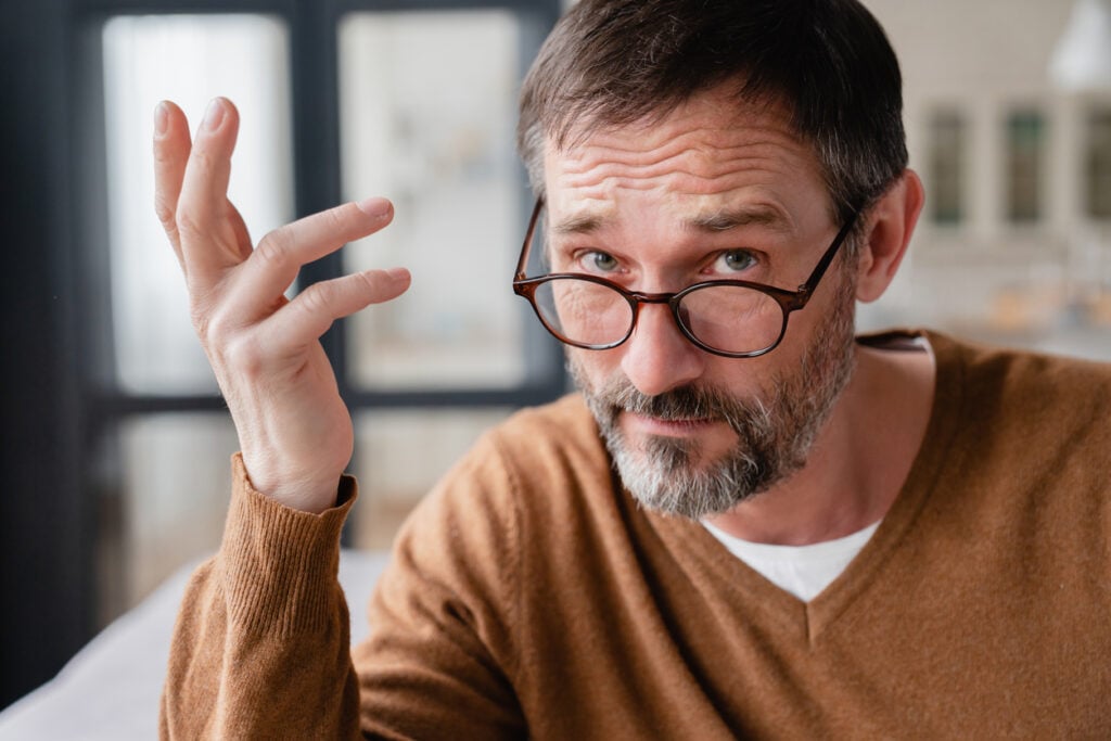Indoor room, bearded man with glasses raising hand, half-body portrait, natural light, documentary style, one person.