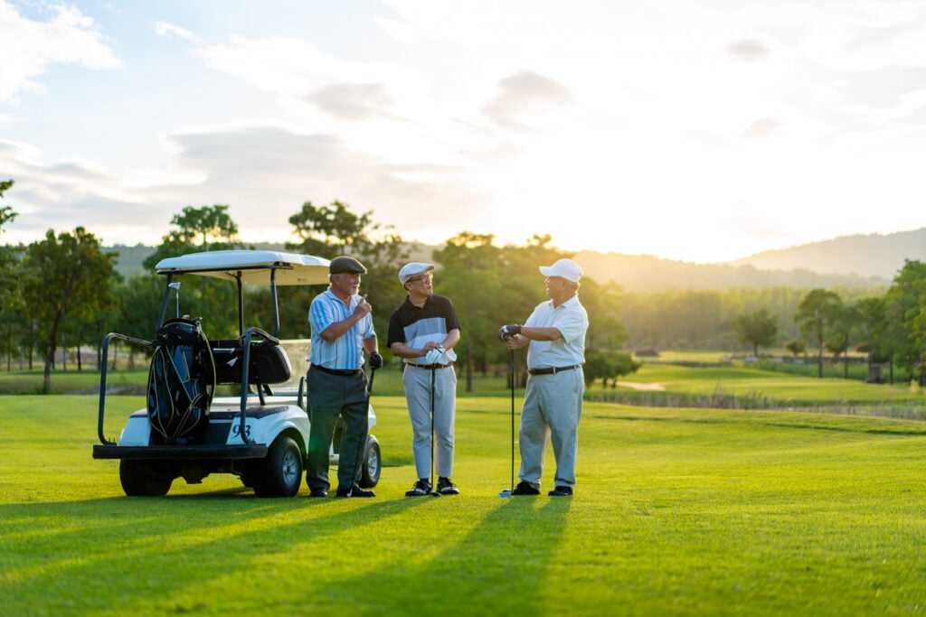 Golf course, three older men with golf cart, one holding club, midday sun, editorial travel photo, people.