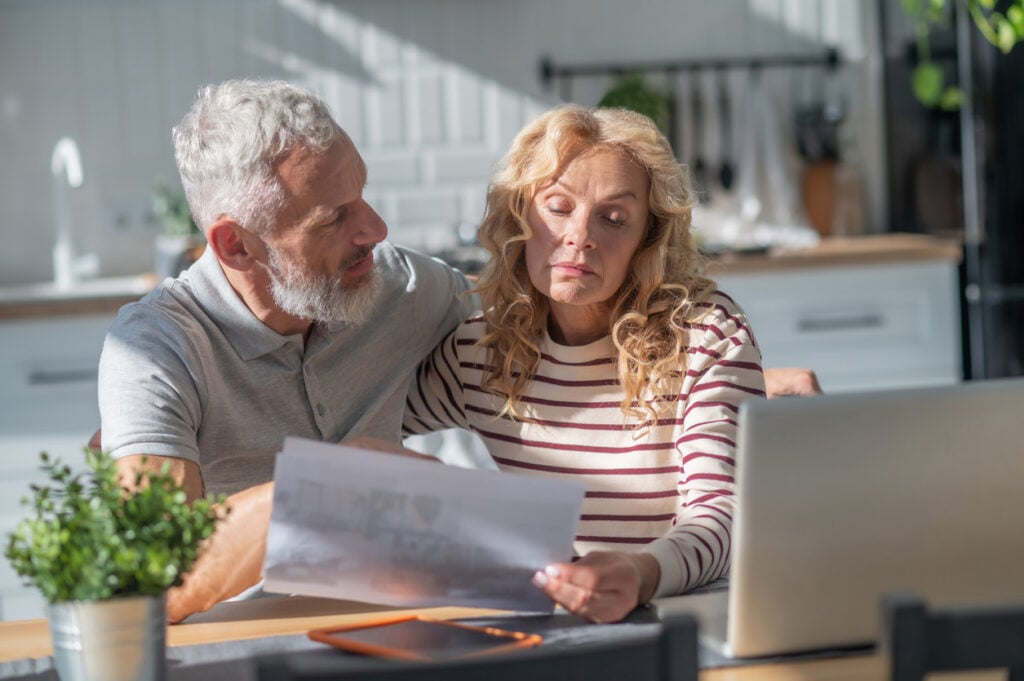 Kitchen table, financial documents in hands, papers and laptop centered, morning sunlight, documentary style, two older adults.