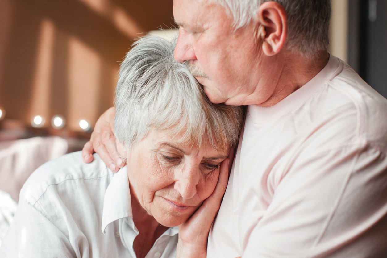 Softly lit room, elder couple embrace, she rests head on his chest, gentle daylight, documentary style, two people.