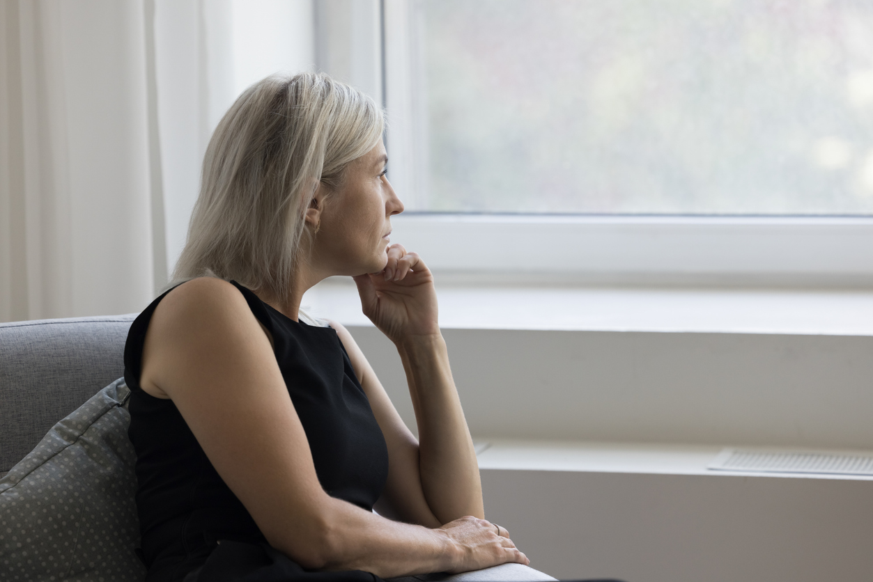 Modern living room, gray-haired woman on sofa, profile view gazing through window, soft daylight, editorial travel photo, people.