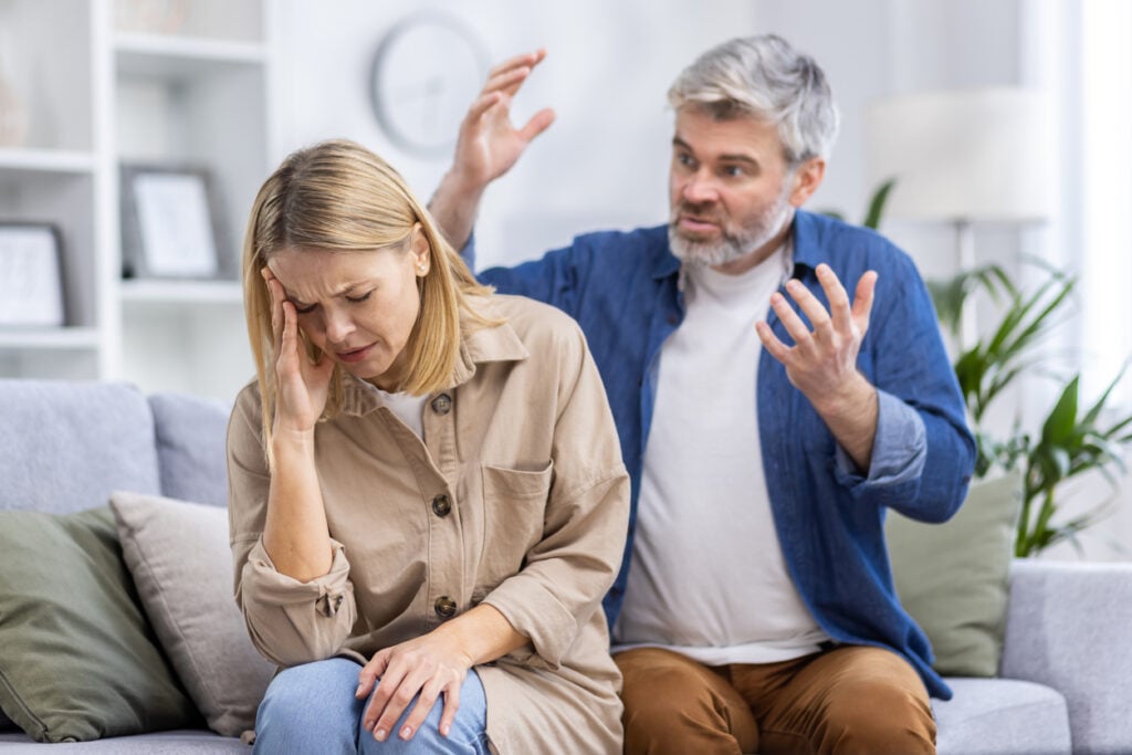Living room, distressed woman foreground, angry man background, natural indoor light, documentary style, two people.