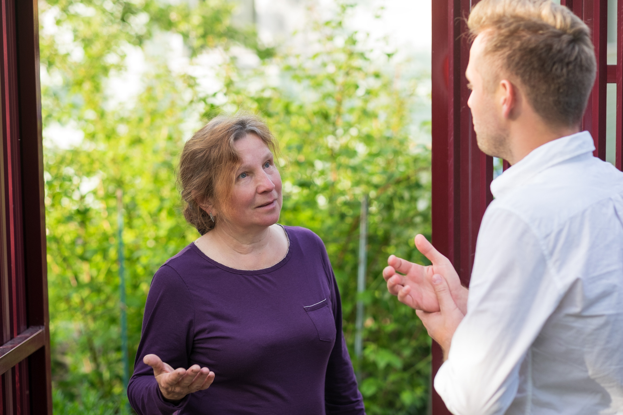 Doorway, woman in purple and man in white converse, bright daylight, documentary style, greenery backdrop, people present.