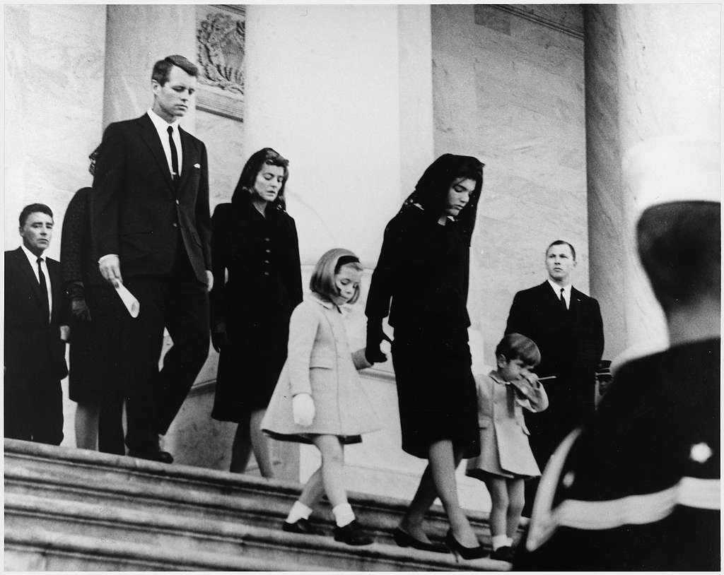 Official building exterior, woman in black leads children down stone steps, daylight, documentary style, several adults.