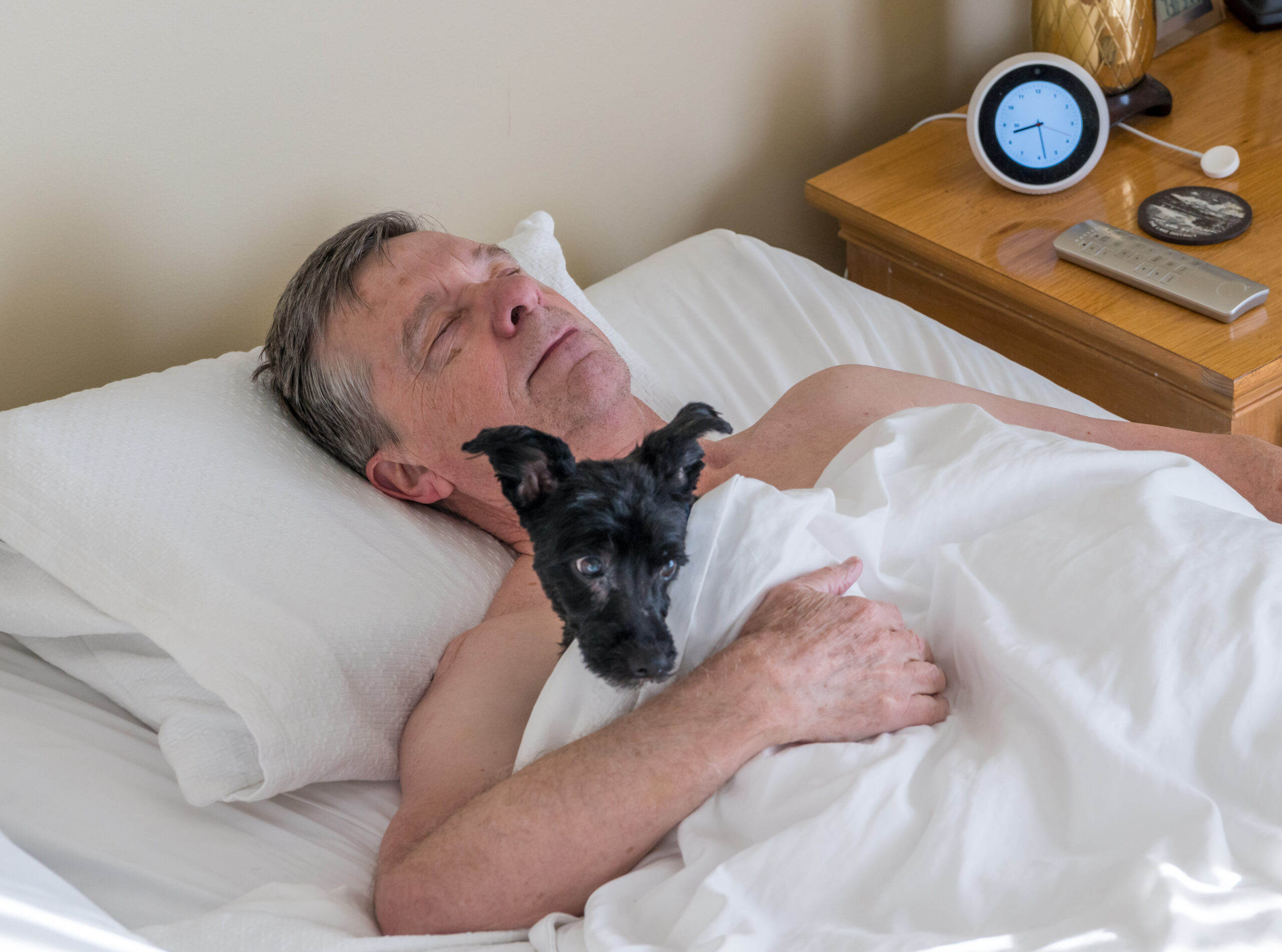 Bedroom, elderly man resting with black dog, nightstand beside bed, morning sunlight streaming in, documentary style, people present.