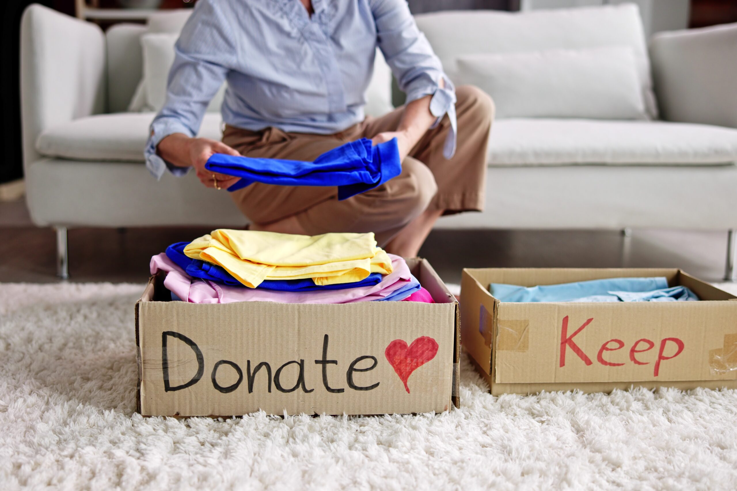 Living room, clothes sorting into Donate and Keep boxes, soft carpet foreground, natural morning light, documentary style, one person.