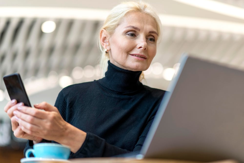 Cafe interior, woman with blonde hair at table, smartphone and laptop open, daylight, editorial travel photo, one person.