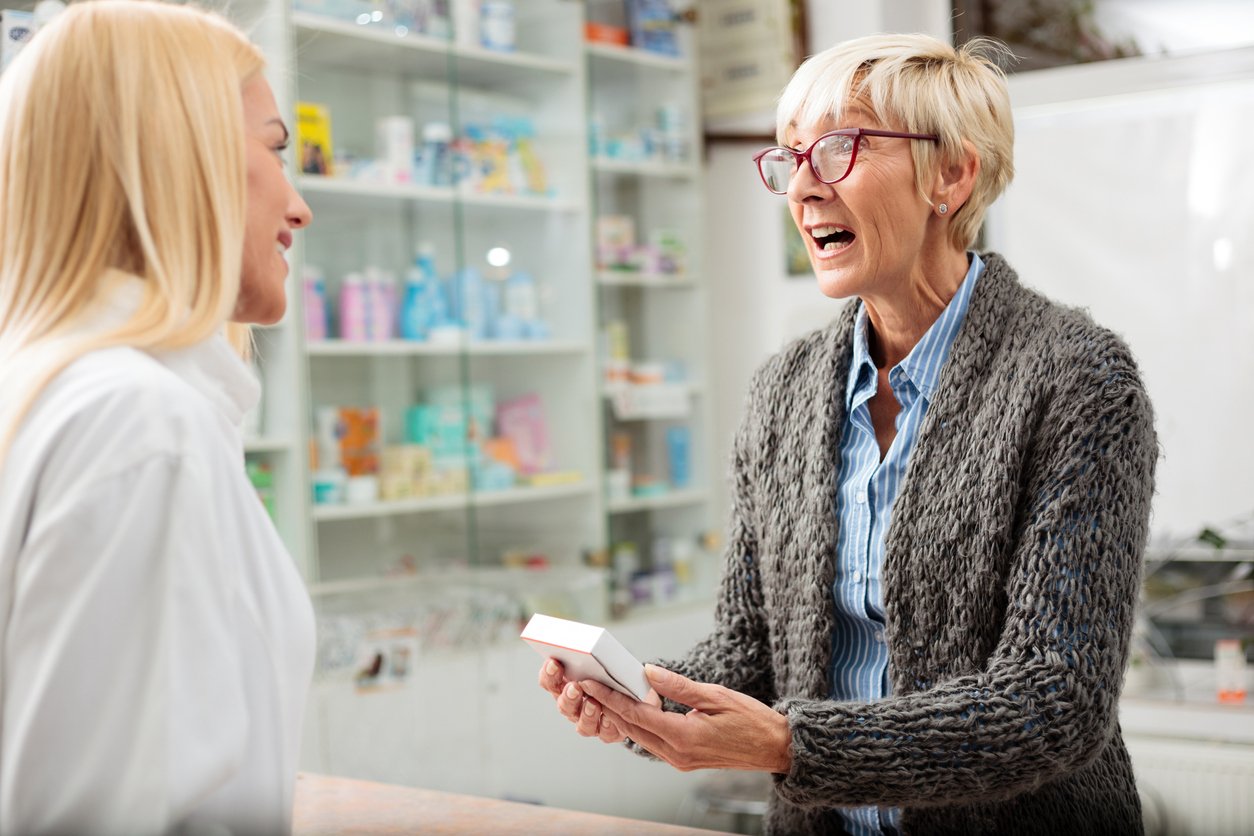 Pharmacy, older woman with glasses holds medicine box, mid-shot, bright indoor light, documentary style, two people.