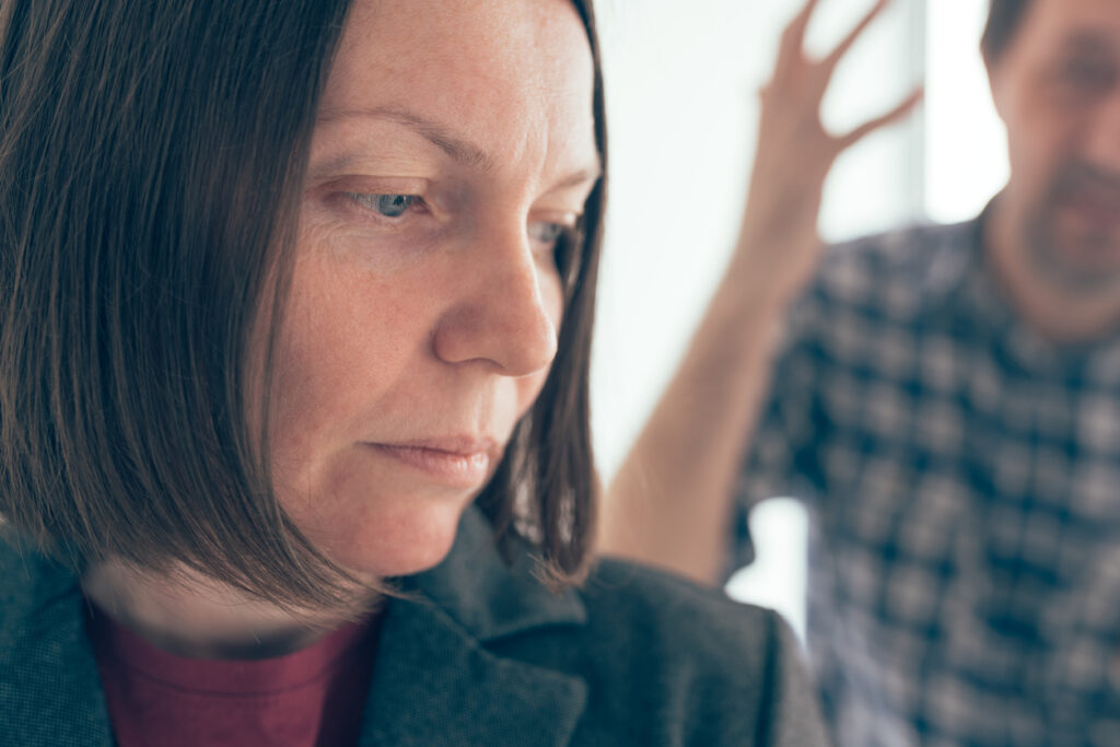 Indoor setting, woman with short brown hair foreground, blurred man background gesturing, soft daylight, documentary style, two people.