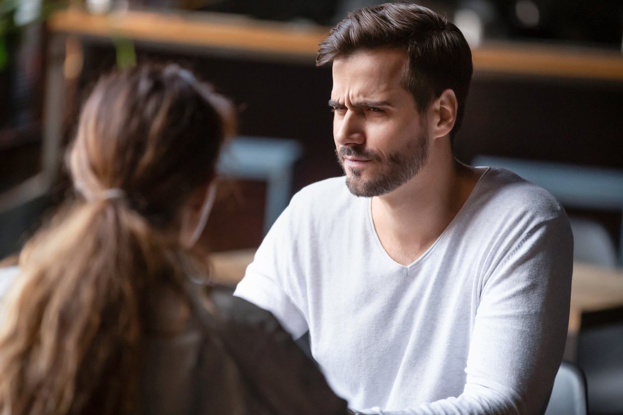 Café interior, man’s skeptical face centered, table foreground, daytime soft light, editorial travel photo, two people.