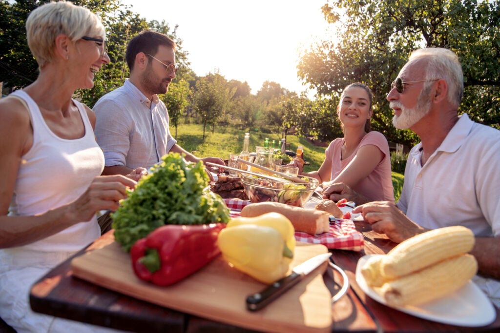 Outdoor picnic area, smiling friends at wooden table, overhead food display, afternoon sunlight filtering, editorial travel photo, four people.