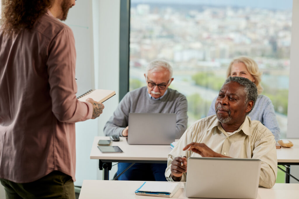 Bright classroom, older adults at desks with laptops, instructor standing, daytime cityscape view, documentary style, people.