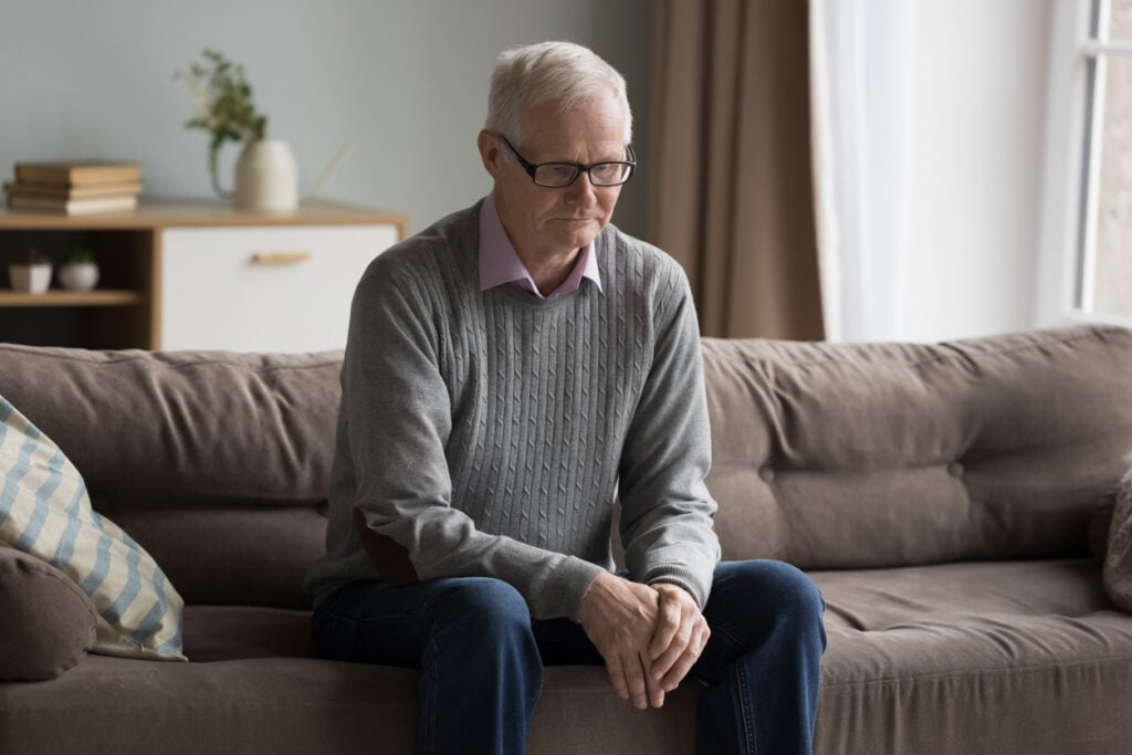 Cozy living room, older man with gray hair, brown couch foreground, soft daylight, documentary style, single person.