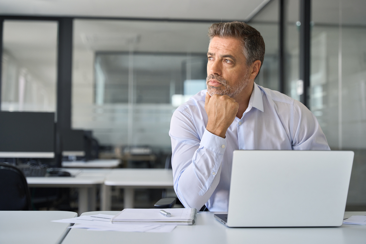 Bright office interior, man in light shirt at desk, laptop and papers arranged, daylight streaming, documentary style, one person.