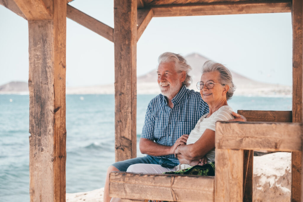 Seaside shelter bench, smiling elderly couple, wide shot, bright daylight, editorial travel photo, people.