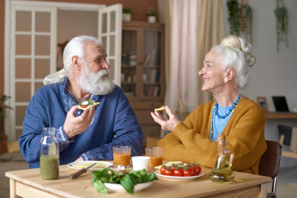 Cozy dining room, elderly couple with sandwiches, food-laden table foreground, daylight glow, editorial travel photo, people.