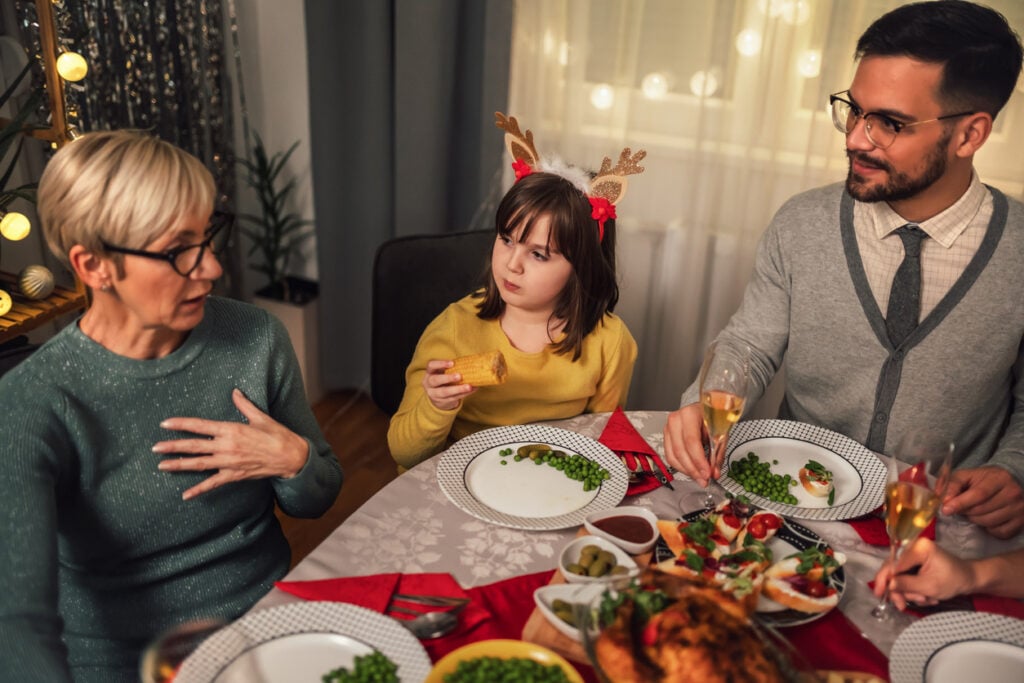 Festive dining room, older woman talking, reindeer-antler headband girl listening, warm evening light, documentary style, people.