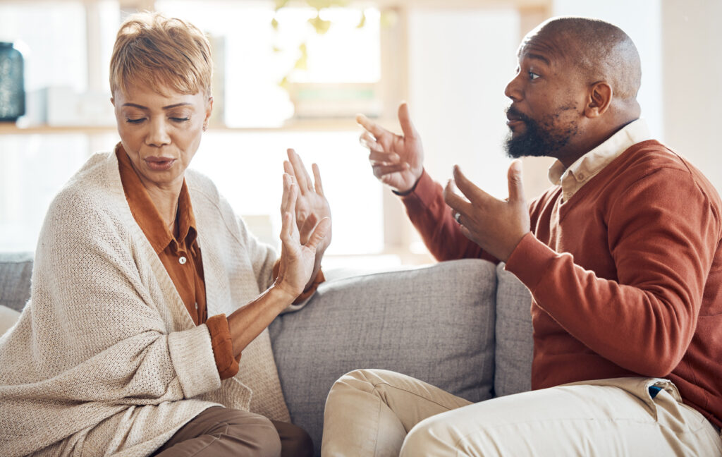 Living room, tense couple on couch, woman defensive, man animated, evening indoor light, documentary style, two people.