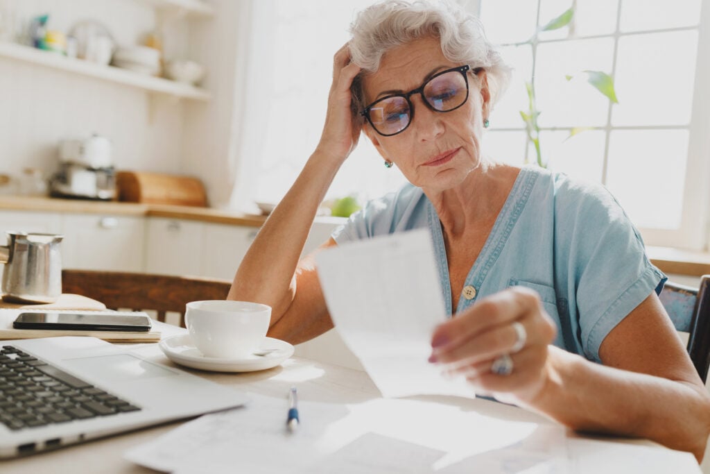 Brightly lit kitchen, concerned older woman, reviewing documents at table, morning light, documentary style, one person.