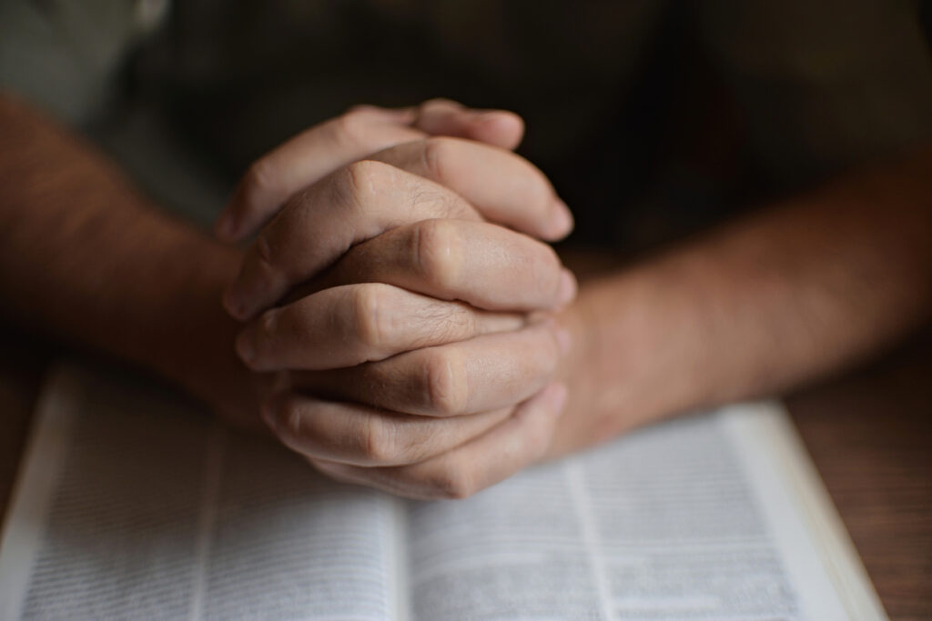 Library table, clasped praying hands over blurred Bible, overhead shot, soft daylight, editorial travel photo, one person.