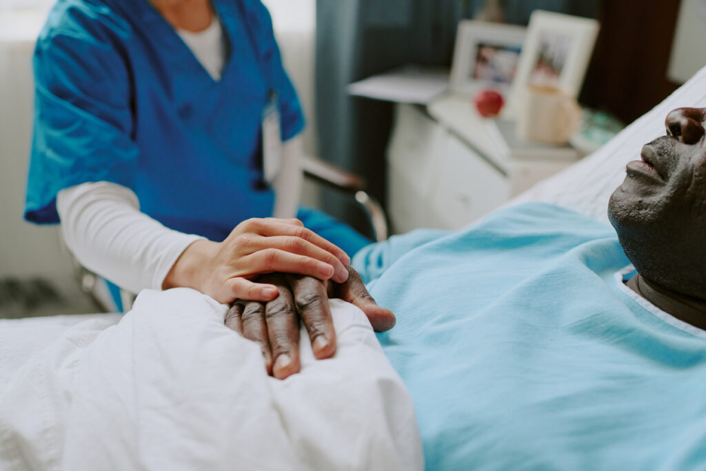 Hospital room, nurse holding patients hand, bedside table in frame, soft daylight, documentary style, two people.