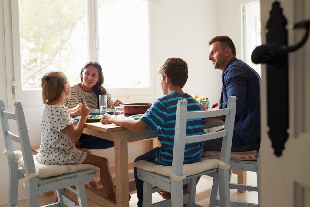 Sunlit dining room, wooden table centerpiece, family gathered for meal, midday light streams in, editorial travel photo, four people.