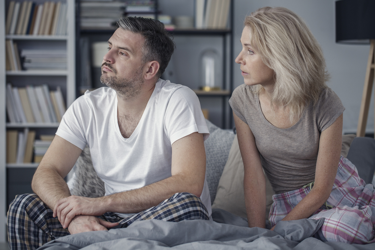 Bedroom, couple in pajamas tense on bed, bookshelves background, soft daytime light, documentary style, two people.