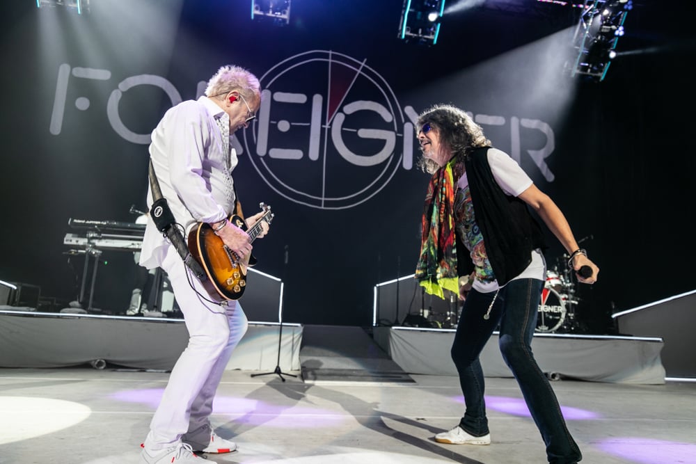 Concert stage, two musicians forefront, band name Foreigner backdrop, bright lights, editorial travel photo, people present.