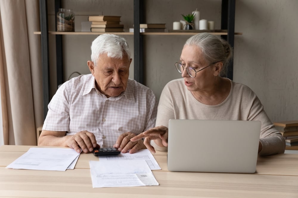 Home office, elderly couple calculating finances, tabletop view, daytime natural light, documentary style, two focused people.