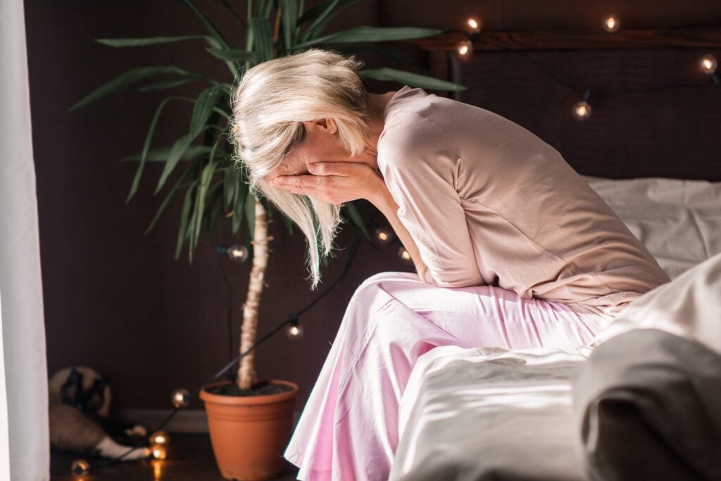 Bedroom interior, distressed woman foreground, seated edge-of-bed, morning soft light, documentary style, one person.