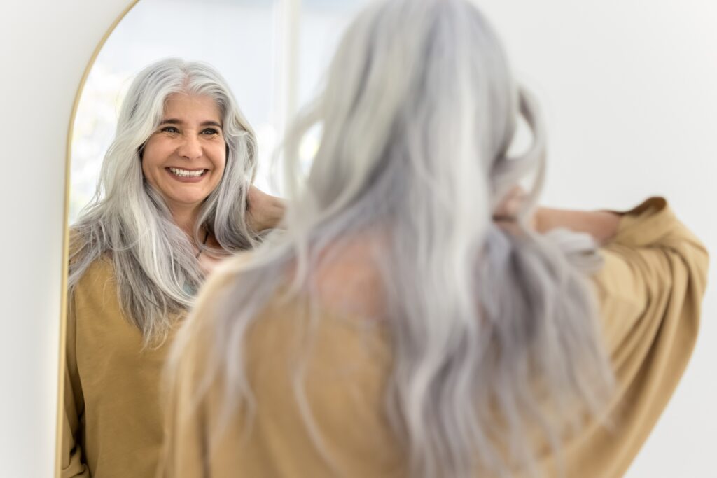 Brightly lit room, gray-haired woman, mirror reflection, soft daylight, editorial travel photo, one person.