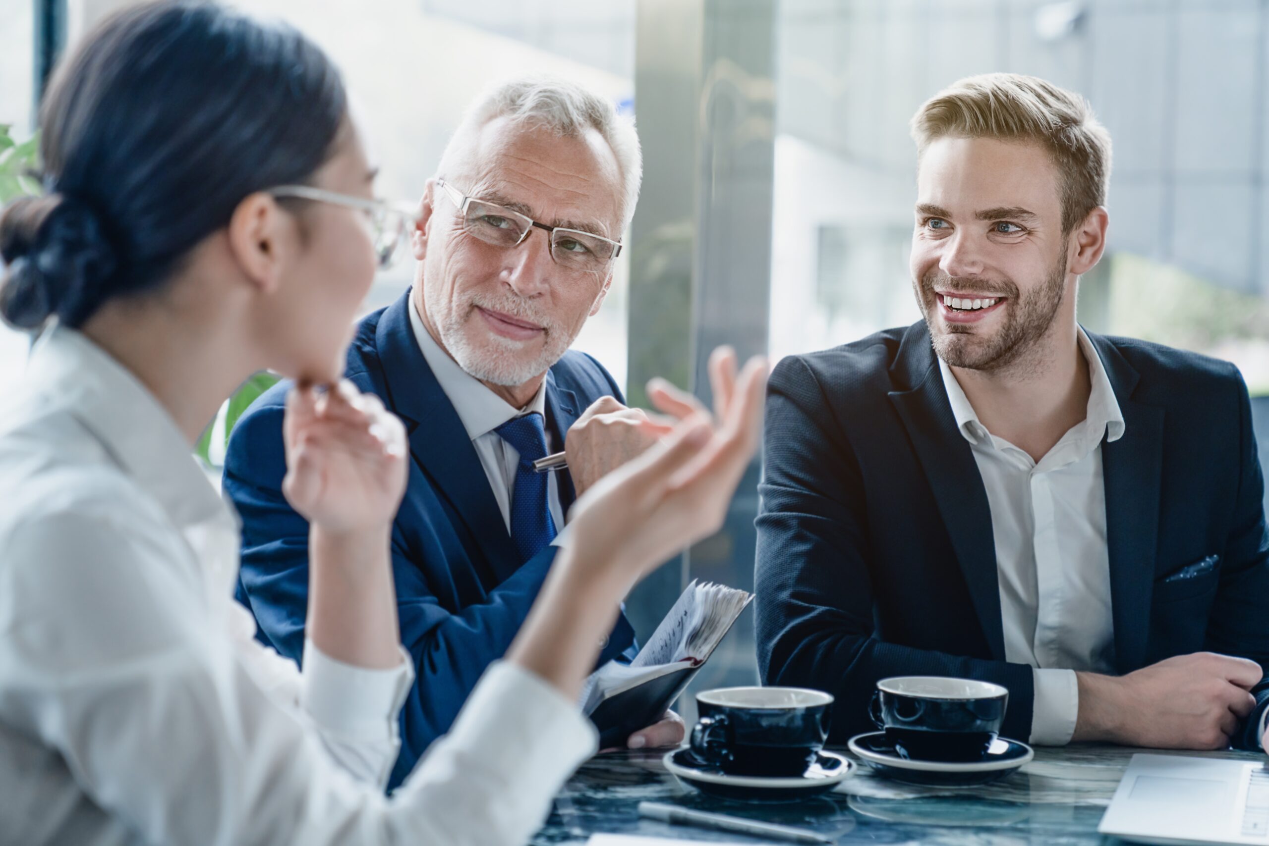 Modern office, three colleagues at table, coffee cups present, bright daylight, documentary style, people engaging conversationally.