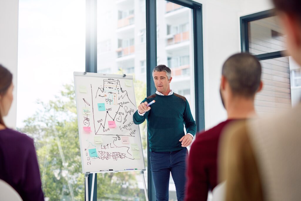 Modern office, whiteboard with charts, presenter at front, daylight streaming in, documentary style meeting scene, three people.