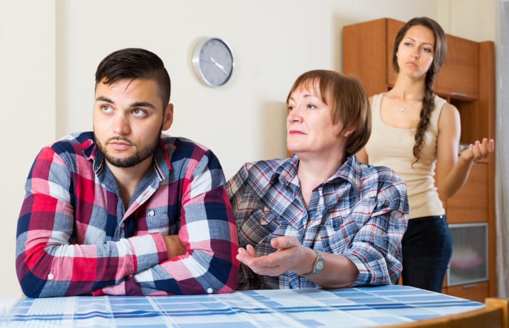 Kitchen interior, tense discussion at table, wide shot, natural light, documentary style, three people visible.