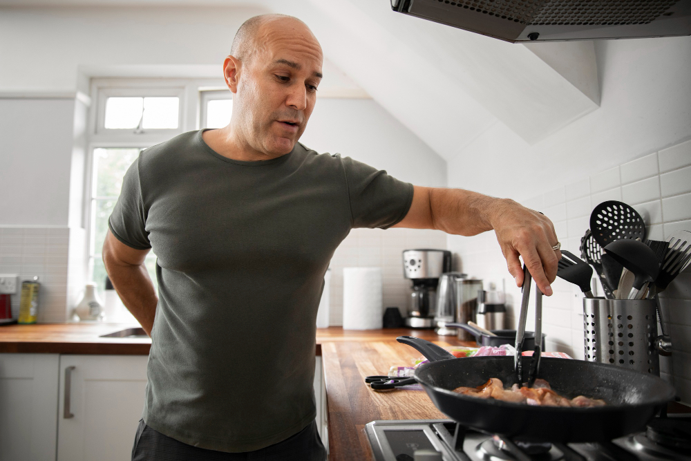 Modern kitchen, man cooking with tongs at stove, countertop utensils, daylight, editorial travel photo, one person present.