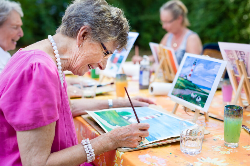 Outdoor art class, elderly woman painting landscape, group around table, midday sunlight, editorial travel photo, people present.